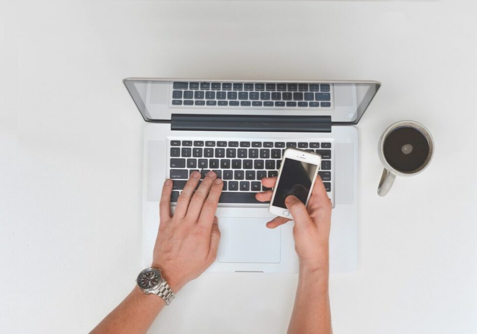 Mans-hands-at-a-laptop-holding-cell-phone-with-coffee-nearby